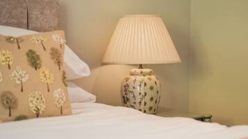 Una cama con un almohadón blanco y uno beige con estampado de árboles, una lámpara blanca con pantalla plisada y una mesita de noche verde.