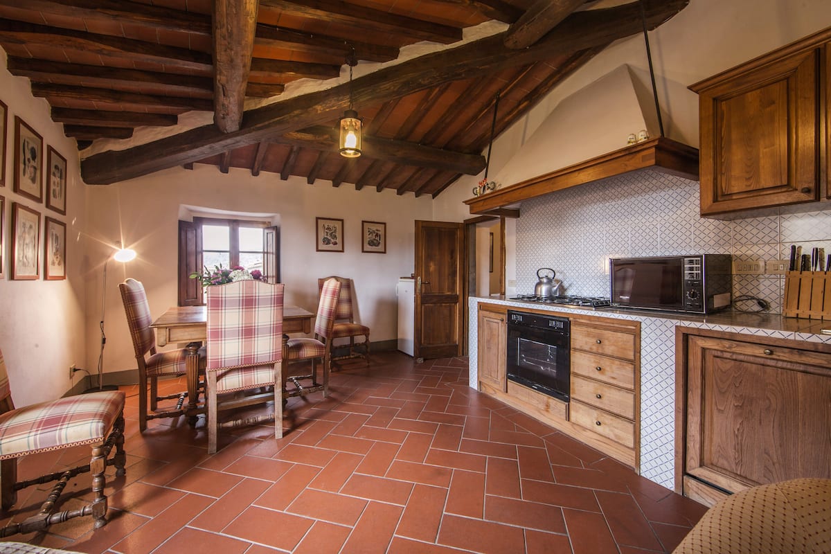 Una cocina con armarios de madera, piso de baldosas y un comedor con sillas a cuadros.