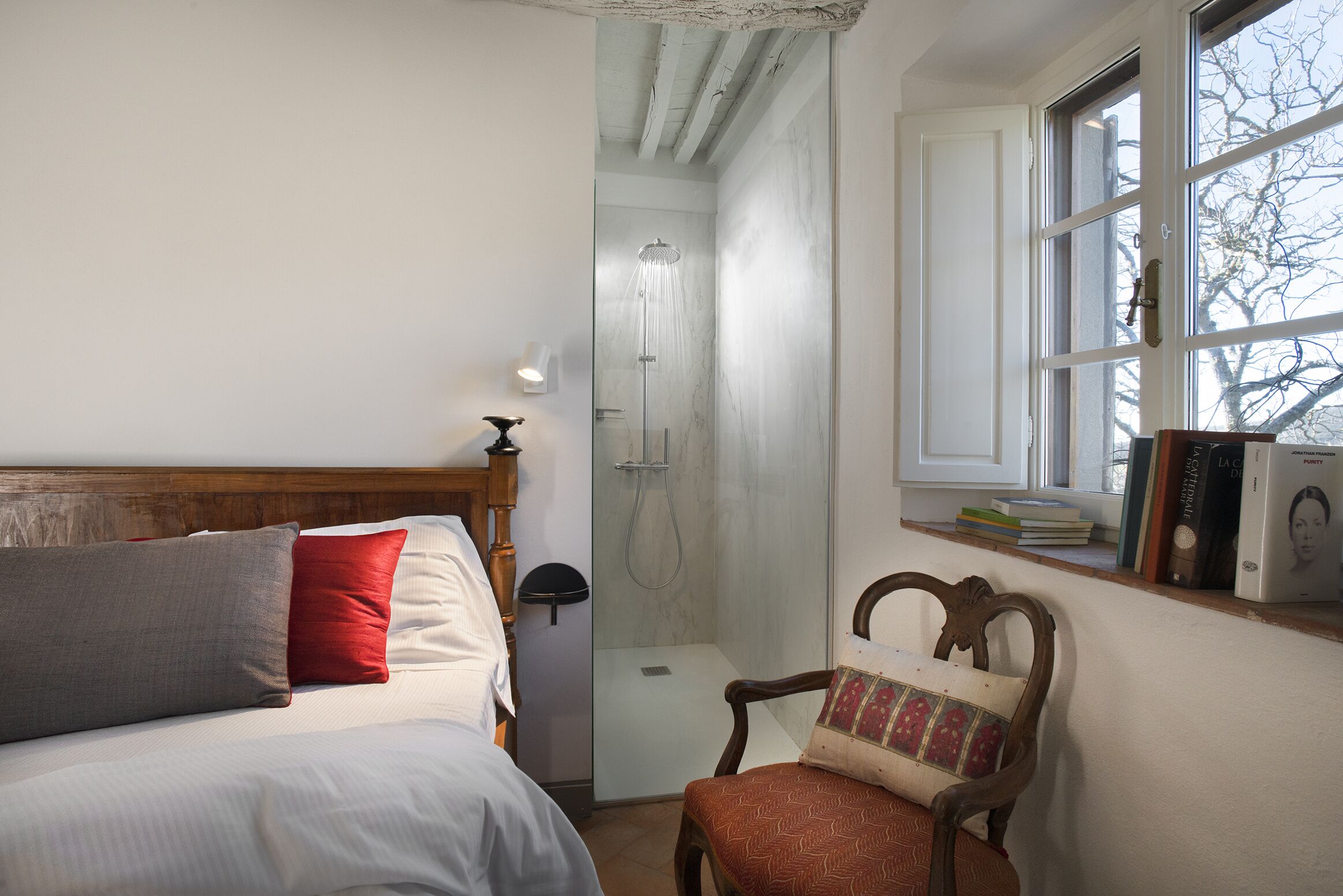 A bedroom with a bed, a chair, a shower, and a window with books on the sill.