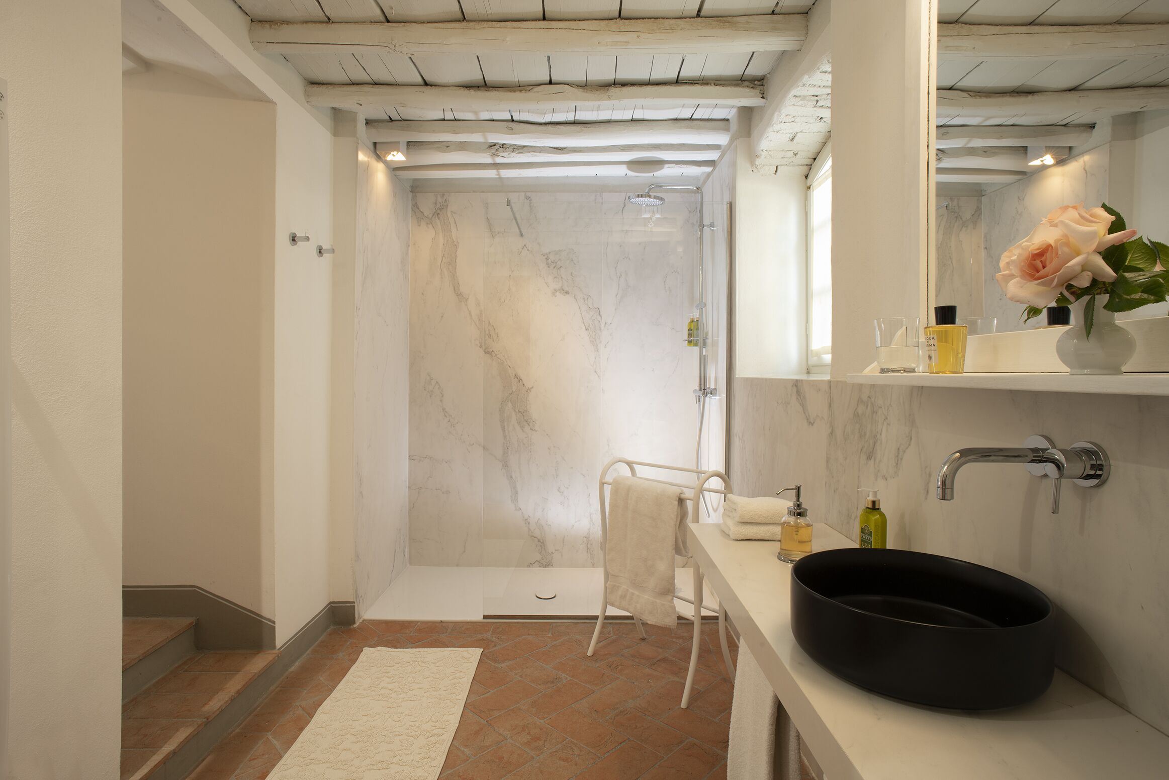 A modern bathroom with a marble shower, a black sink, and a white towel on a chair.