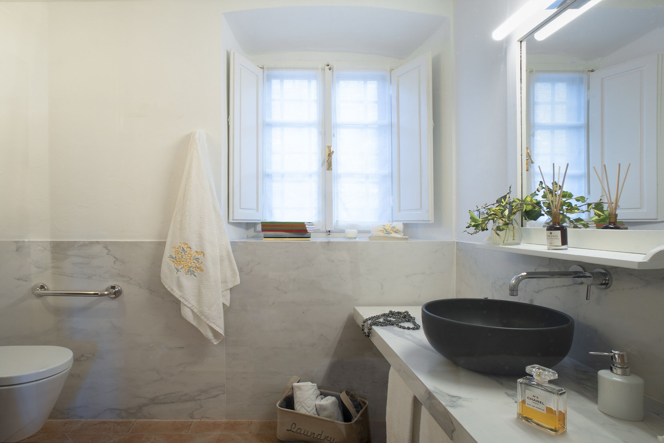 Un baño con pared de mármol, un lavabo tipo cuenco negro, un inodoro blanco, un perchero y una ventana con vistas al exterior.