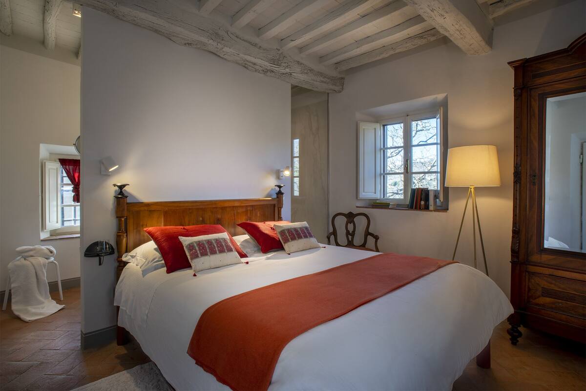 Una cama bien hecha con cabecera de madera, una lámpara de pie y una ventana con vista a árboles.