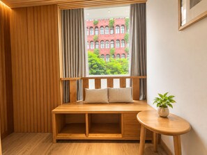 Un banc en bois doté d’un siège et d’un dossier rembourrés, une petite table sur laquelle est posée une plante en pot, et une vue sur un bâtiment rouge, visible à travers une fenêtre ornée de rideaux.