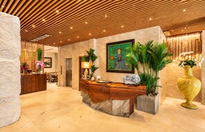 Zona de recepción con un mostrador de madera, piso de mármol, un cuadro grande y plantas en macetas.