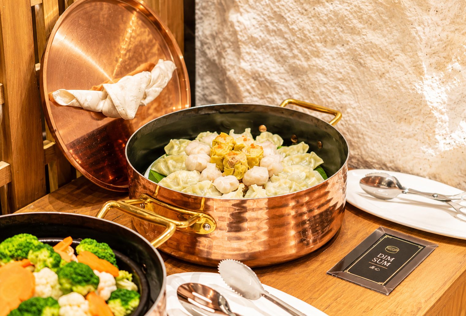 A copper pot with dumplings and vegetables on a wooden table, accompanied by a spoon and a menu.