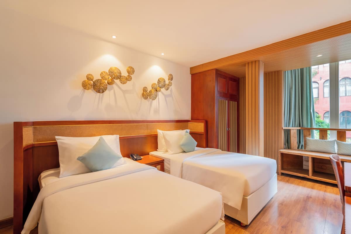 Une chambre d’hôtel avec deux lits, une tête de lit en bois, un bureau avec une chaise et une vue sur un immeuble à travers de grandes fenêtres.
