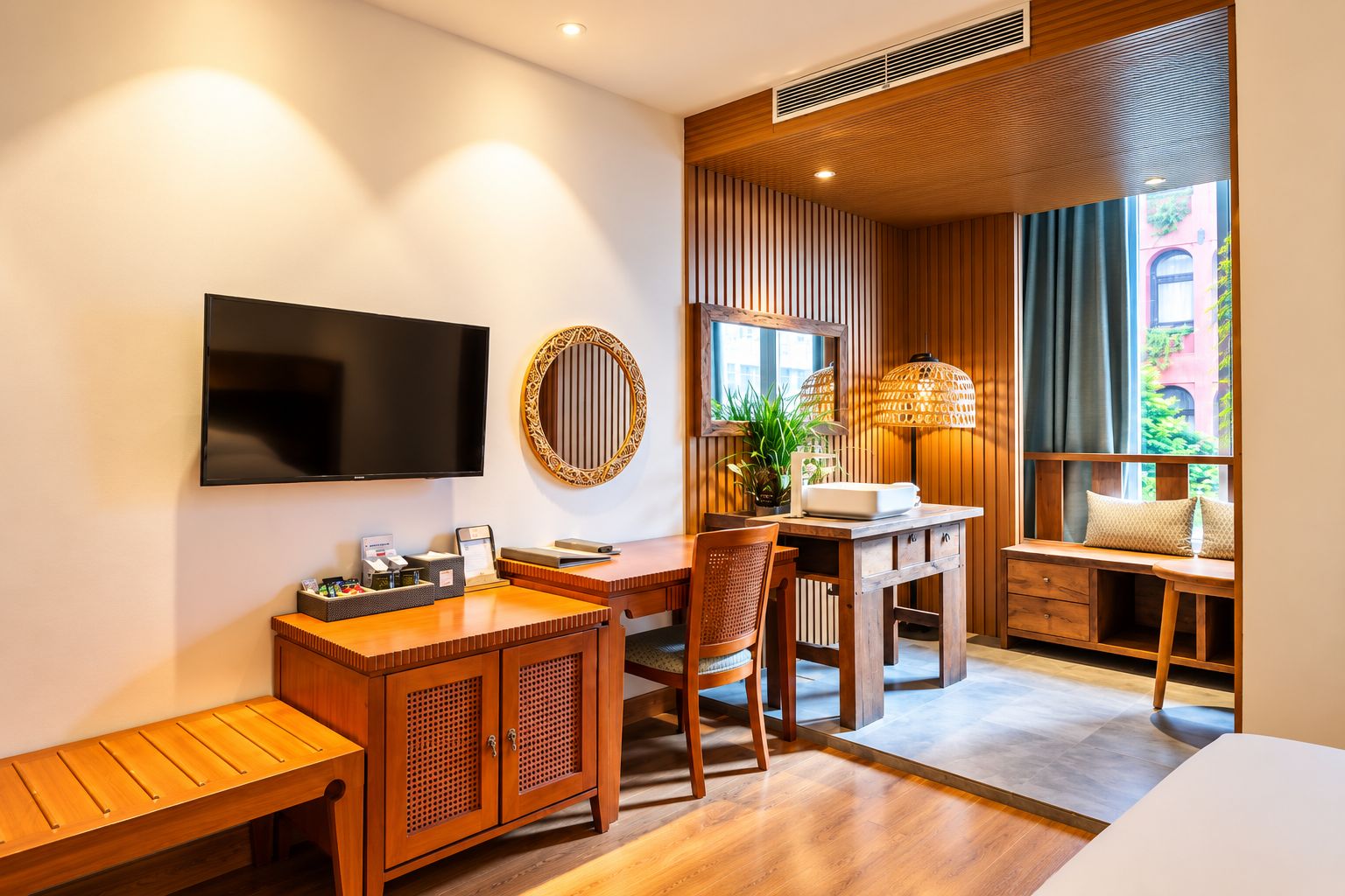 A hotel room with a wooden desk, a television, a round mirror, a window seat with a view, and a ceiling vent.