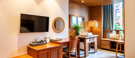 A hotel room with a wooden desk, a television, a round mirror, a window seat with a view, and a ceiling vent.