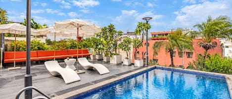 A swimming pool with lounge chairs and umbrellas, surrounded by greenery and buildings.