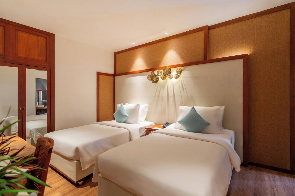 Une chambre d’hôtel avec deux lits, des têtes de lit en bois, un miroir et une plante.