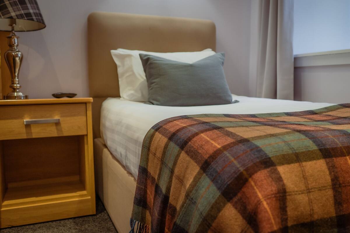 A neatly made bed with a plaid blanket, a bedside lamp, and a wooden nightstand.