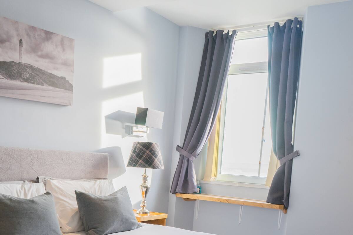 A bedroom with a bed, pillows, a lamp, a window with curtains, and a painting on the wall.