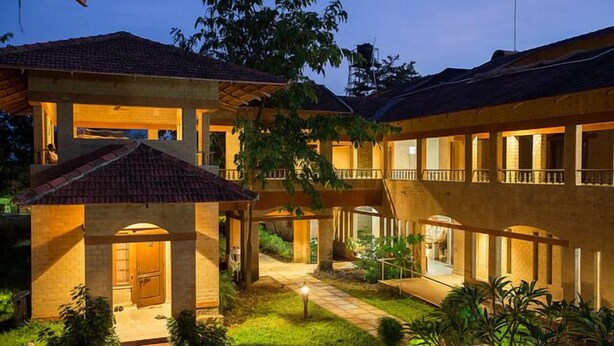 A multi-story hotel with a courtyard, featuring a central open space with a pathway, surrounded by lush greenery and a well-lit entrance. - Govardhan Eco Village (Vada)