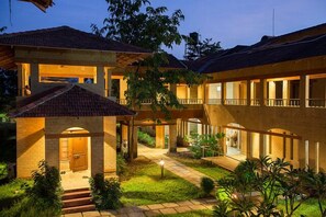 A multi-story hotel with a courtyard, featuring a central open space with a pathway, surrounded by lush greenery and a well-lit entrance. - Govardhan Eco Village (Vada)