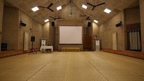 A large, well-lit room with a stage, ceiling fans, and a projector screen. - Govardhan Eco Village (Vada)