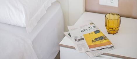A neatly made bed with white linens, a bedside table with a lamp, a glass of water, and a newspaper.