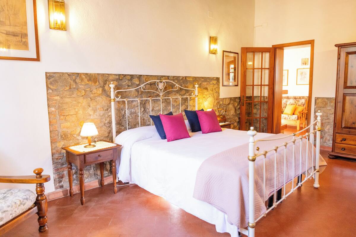Une chambre avec un mur en pierre, un cadre de lit en métal blanc, une table de chevet avec une lampe et une armoire en bois.