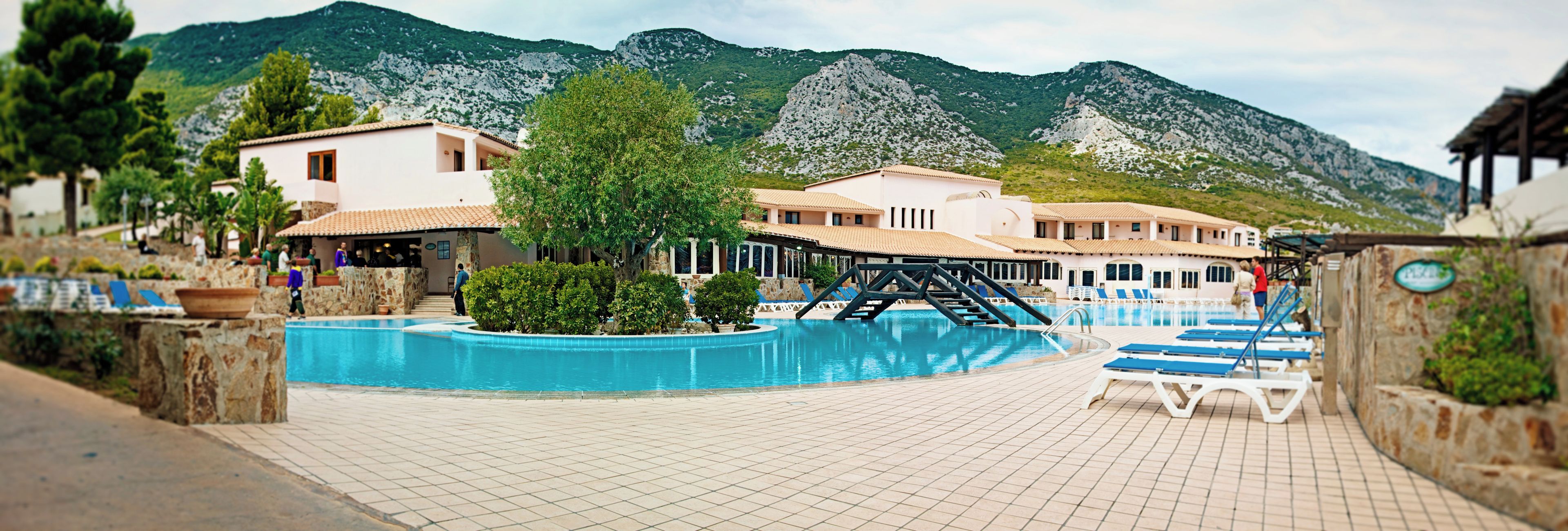 Une piscine avec une vue sur les montagnes, un espace couvert avec des chaises longues et un bâtiment doté d’un toit en tuiles.