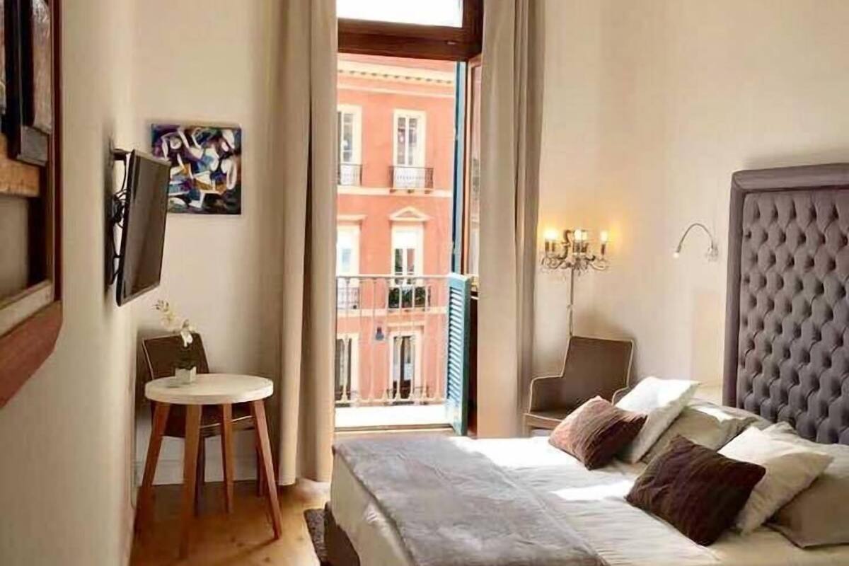 Ein Hotelzimmer mit Bett, Nachttisch, Sessel und einem kleinen Tisch mit Stuhl. Ein Fenster mit Vorhängen, ein Bild an der Wand und Blick auf Gebäude im Freien.