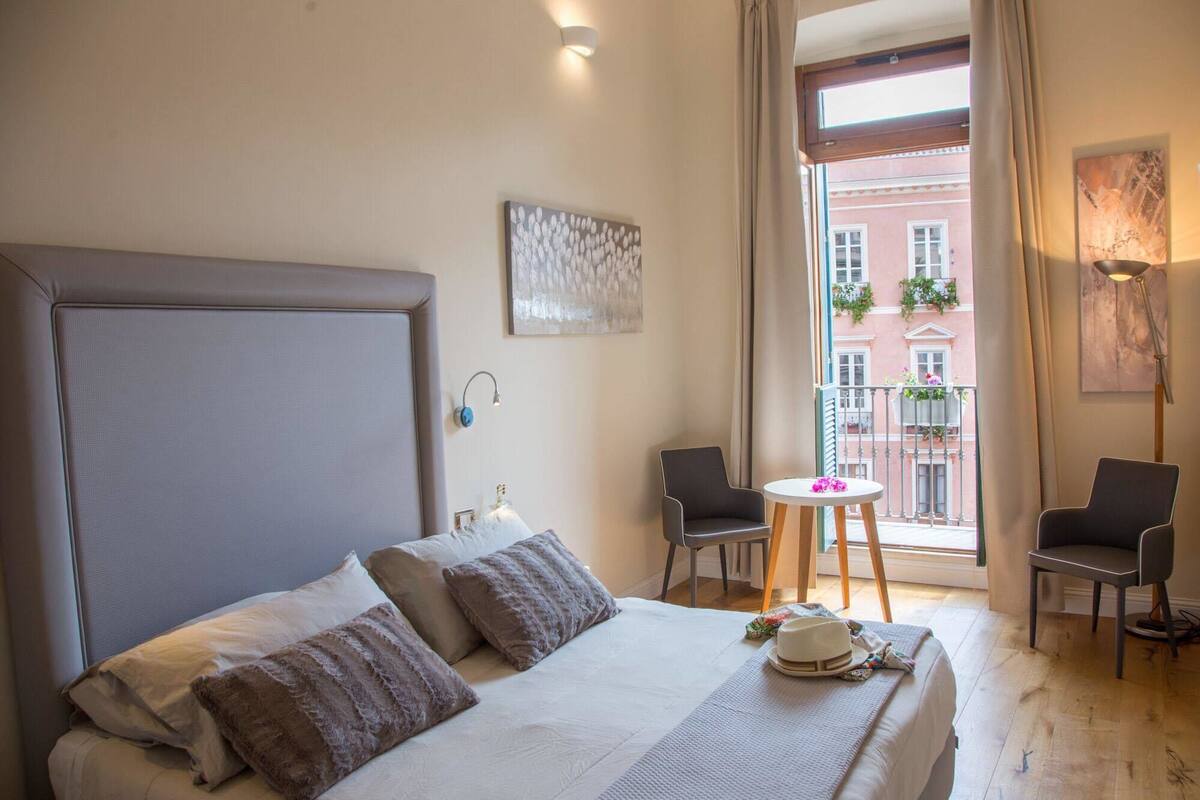 Ein Hotelzimmer mit einem großen Bett, zwei Stühlen, einem kleinen Tisch und einem Fenster mit Blick auf Gebäude.