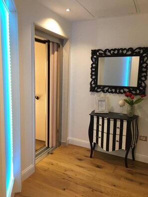 Un pasillo con suelo de madera, una consola a rayas negras y blancas, un espejo decorativo y una puerta con una tira de luz.