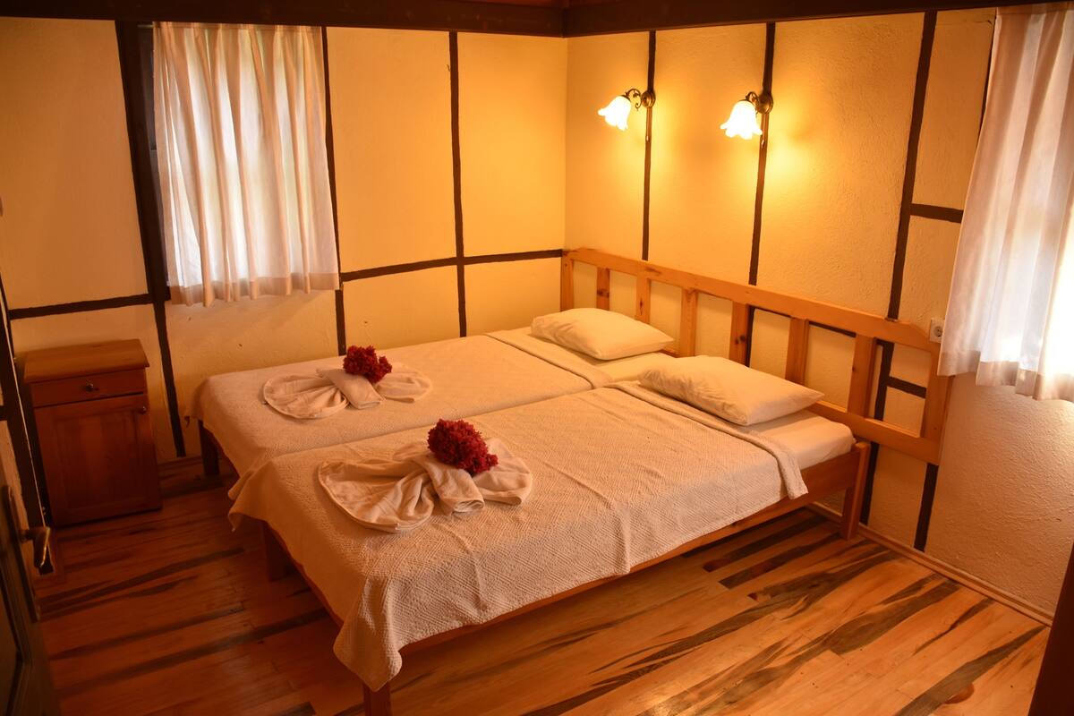 Une chambre avec un lit en bois, du linge de lit blanc et une tête de lit en bois. On y trouve deux lampes fixées au mur et une petite table de chevet en bois.