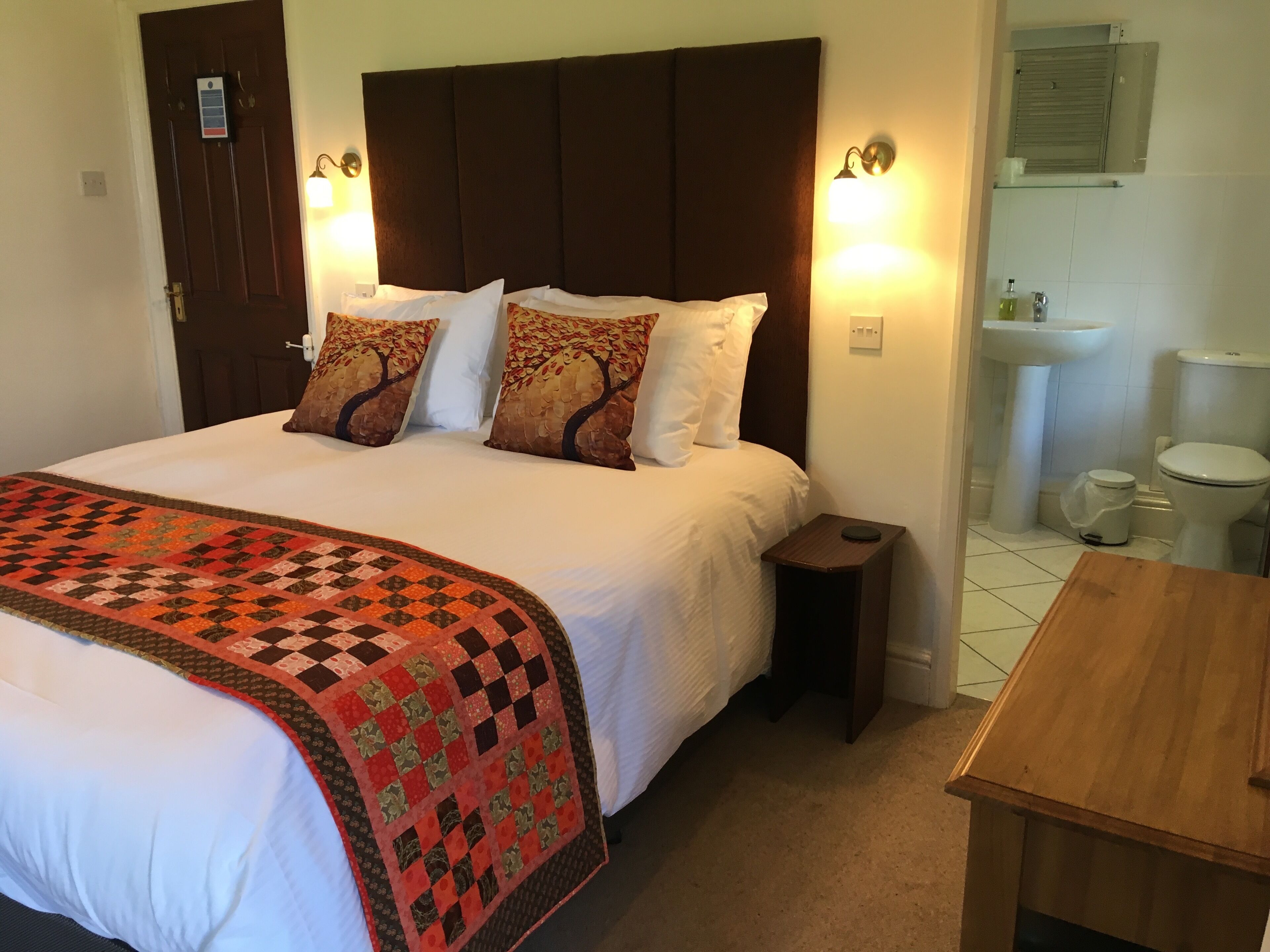 A hotel room with a large bed, a wooden nightstand, a small table, and a bathroom visible through an open door.