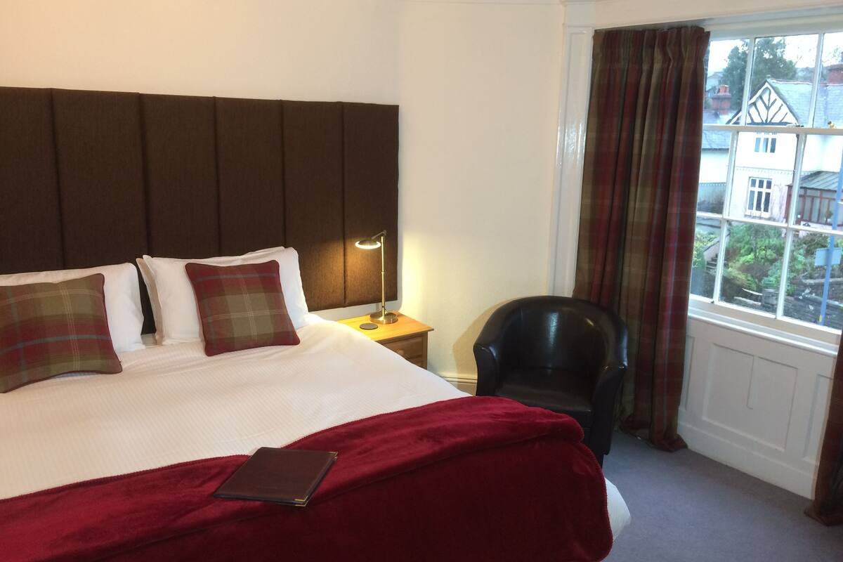 A hotel room with a large bed, a bedside lamp, a chair, and a window with curtains.
