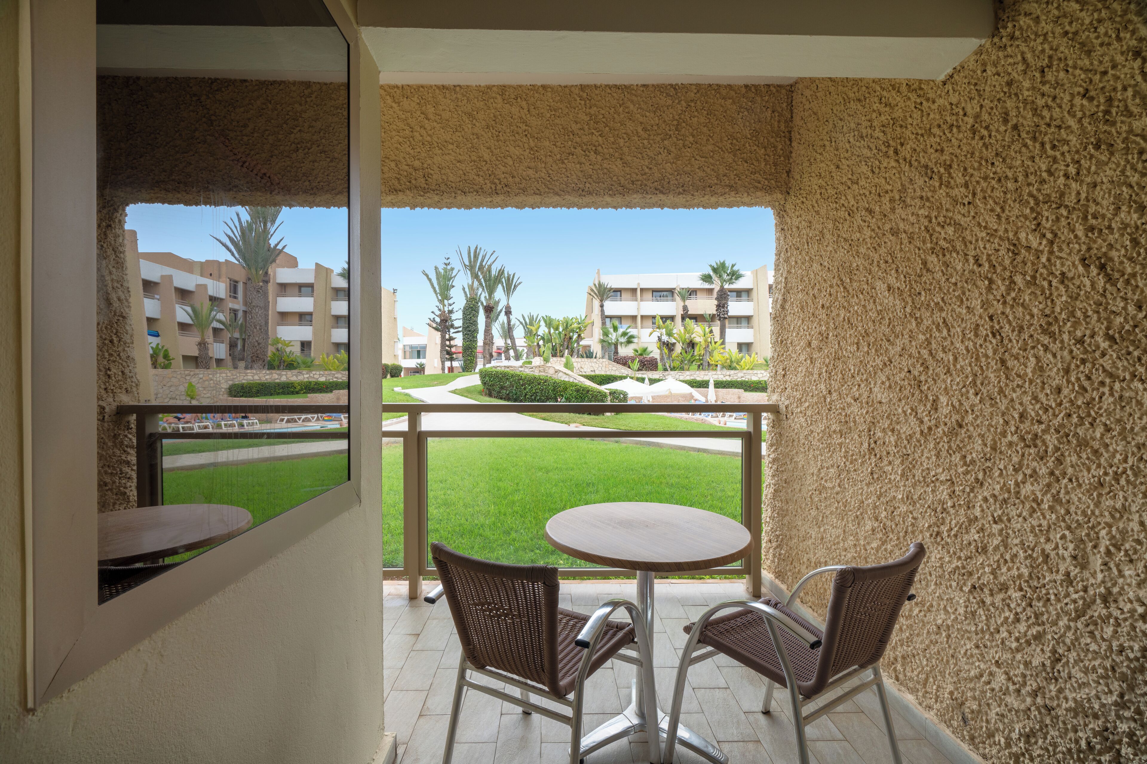 Een balkon met twee rieten stoelen en een kleine ronde tafel, met uitzicht op een verzorgd gazon en gebouwen.