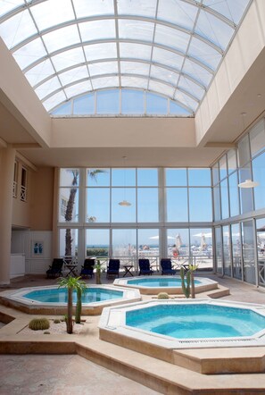 Amplia zona de piscina interior con varias piscinas, rodeada de sillas de descanso y palmeras, bajo un techo de cristal con vista a la playa.