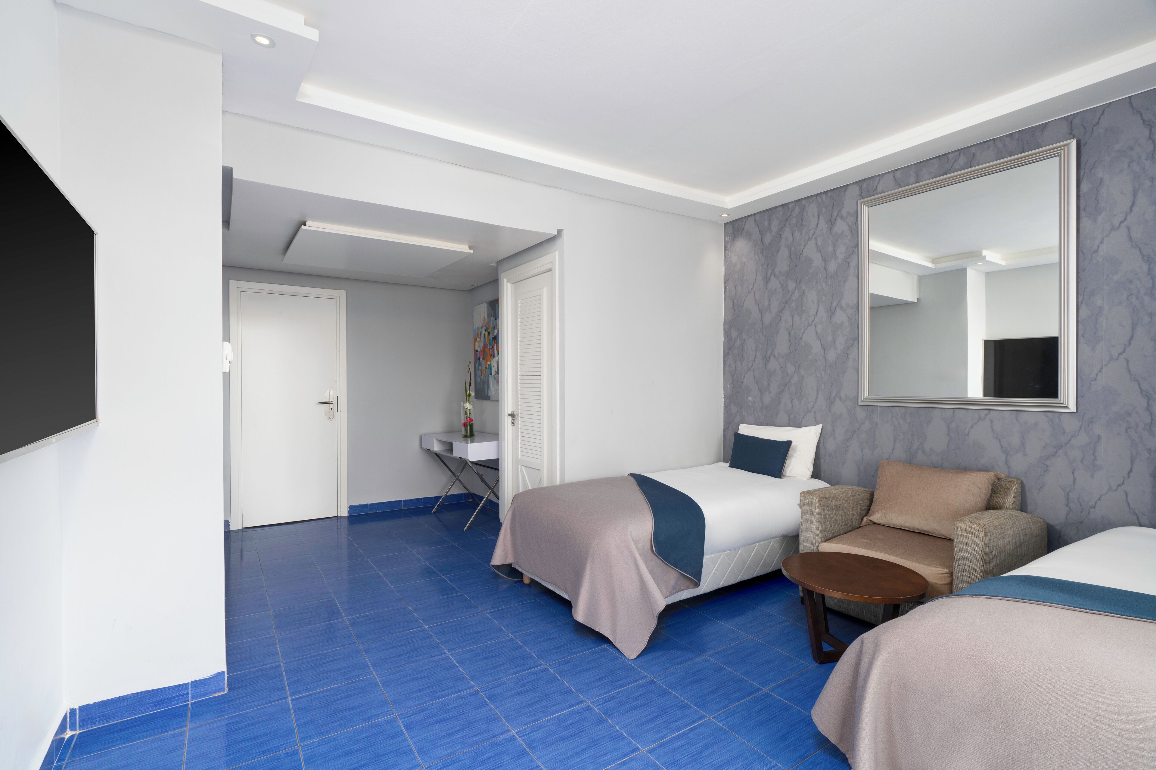 Habitación de hotel con dos camas, un sofá, una mesita, un espejo y un baño con ducha e inodoro.