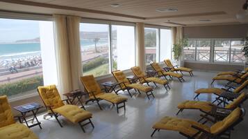 A sunlit room with yellow cushioned chairs arranged in rows, large windows offering a view of the beach and ocean, and a potted plant near the windows.