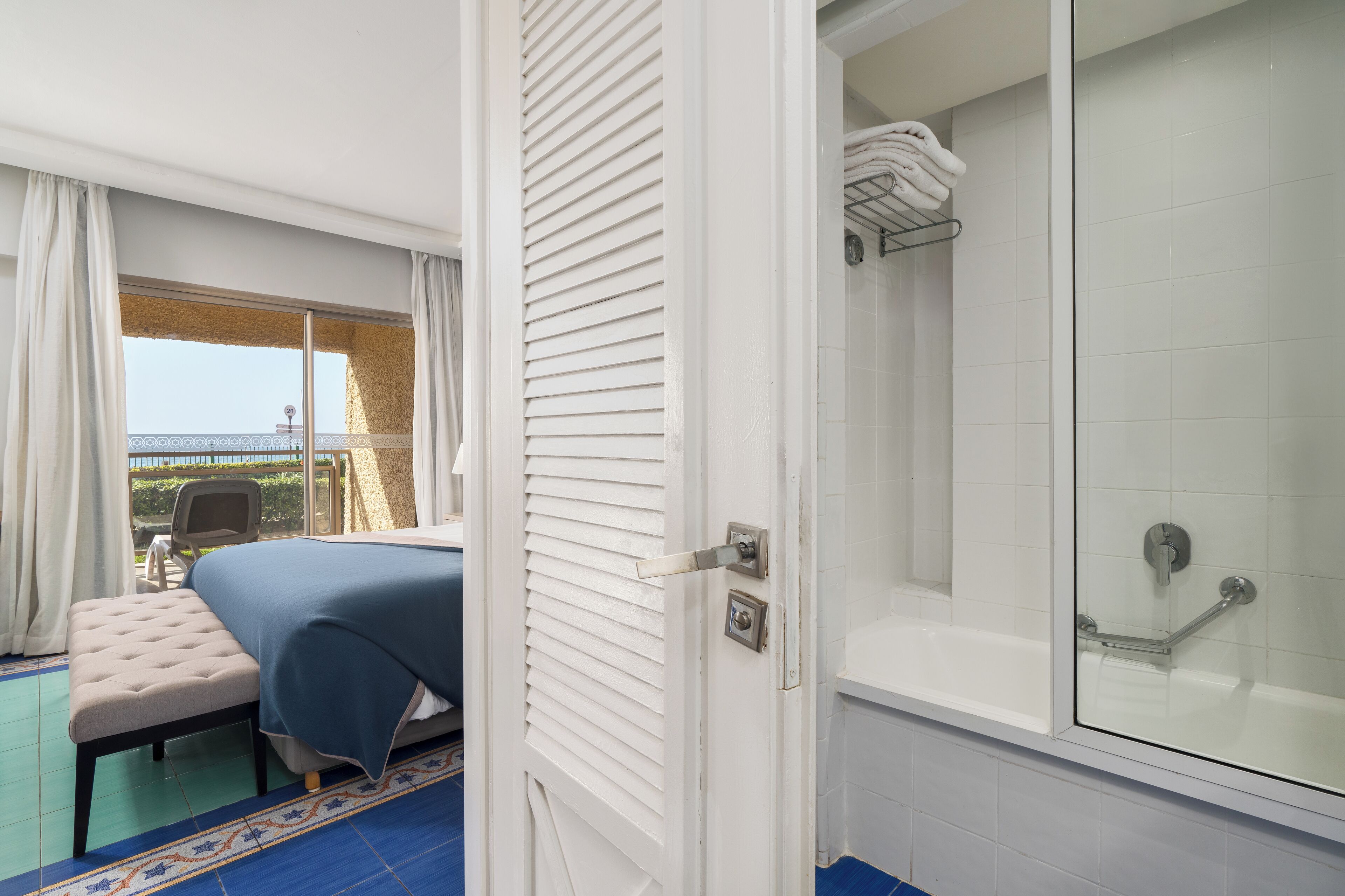 A hotel room with a bed, a bench, a shower, and a view of the outside.