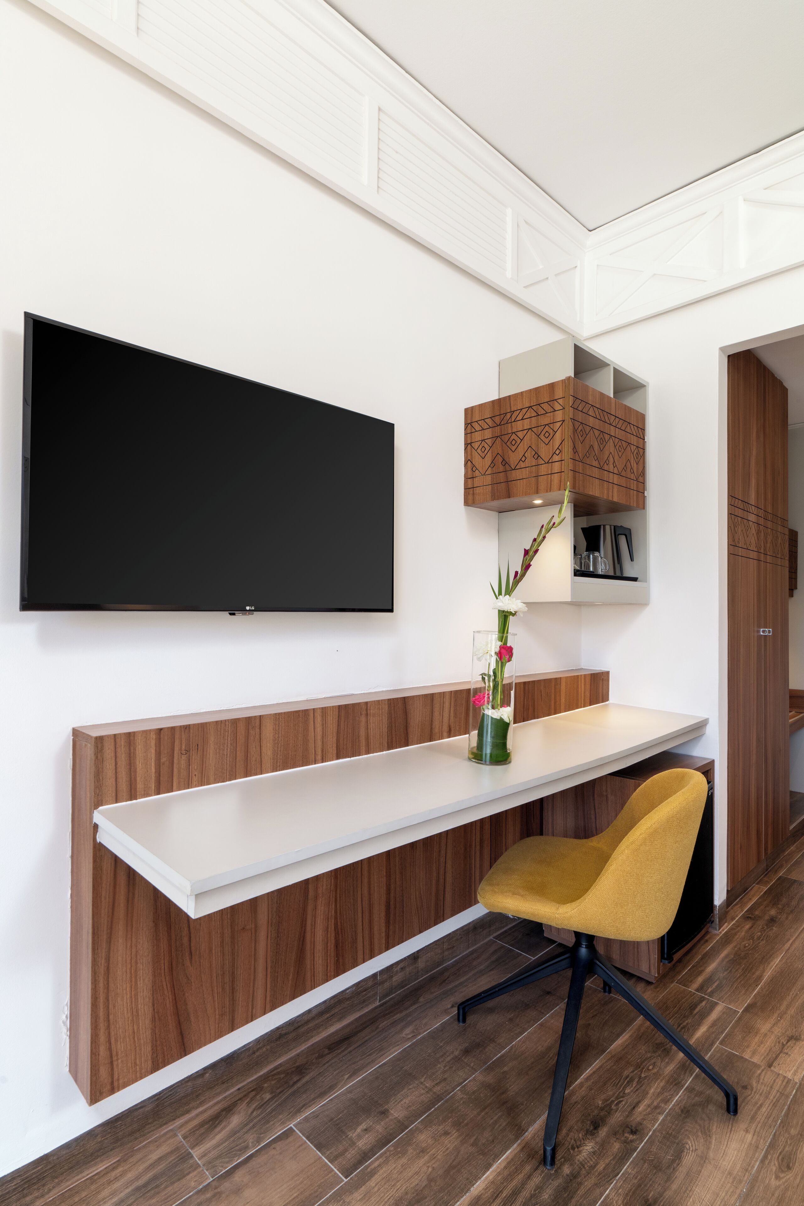A modern office space with a flat-screen TV mounted on the wall, a wooden desk with a white surface, a yellow office chair, and a decorative vase with flowers.