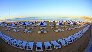 A beach with rows of white lounge chairs and blue umbrellas, arranged in a semi-circle facing the ocean. - Dunes d'Or Ocean Club (Agadir)