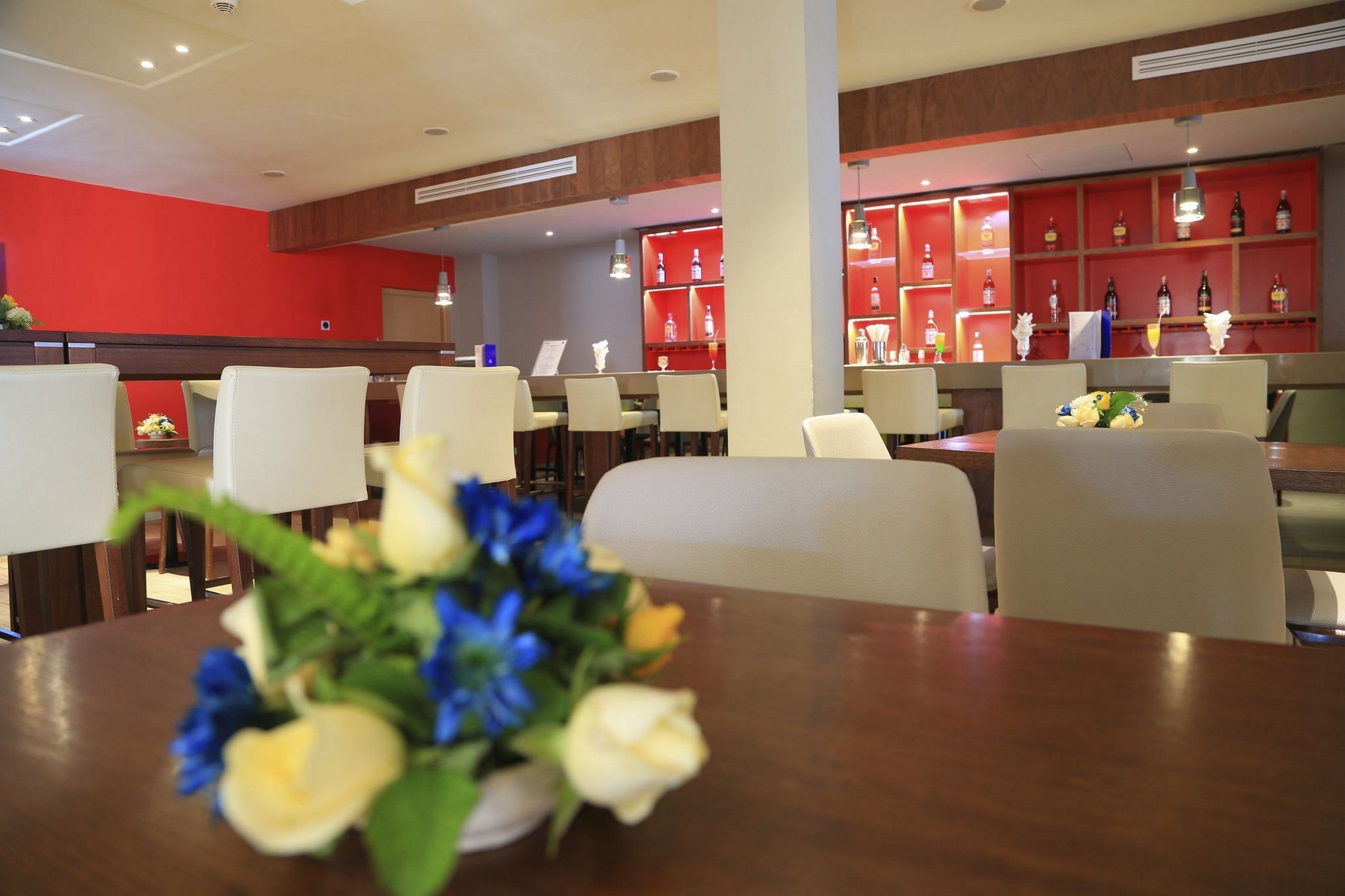 A hotel lobby with a red wall, a bar area with shelves of bottles, and a dining area with tables and chairs.