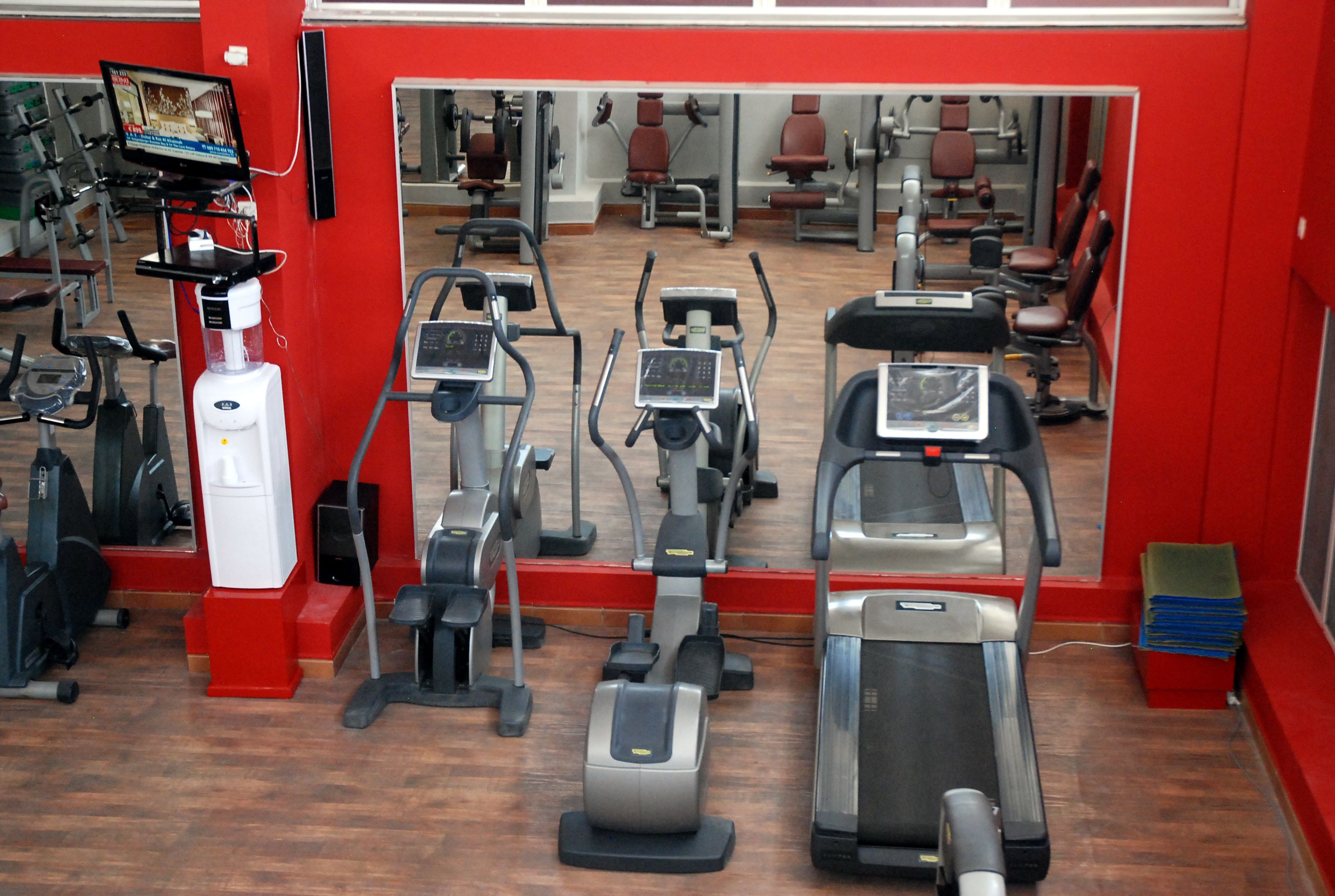 A gym with various exercise machines, a TV screen, and a mirror reflecting the equipment.