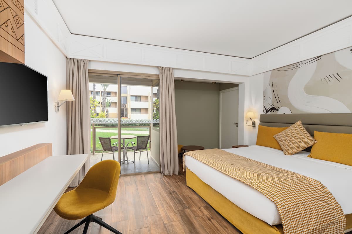 Ein Hotelzimmer mit Bett, Schreibtisch, Stuhl, Fernseher und einem Balkon mit Blick auf ein Gebäude und Grünflächen.