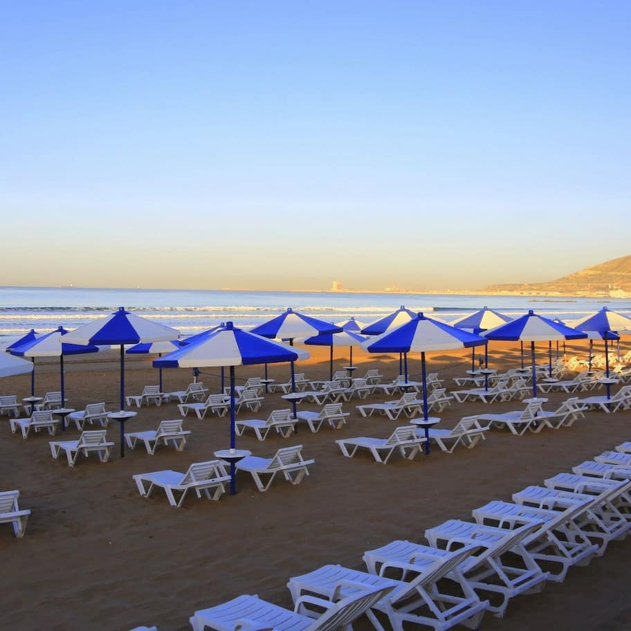 A beach with rows of white lounge chairs and blue umbrellas.