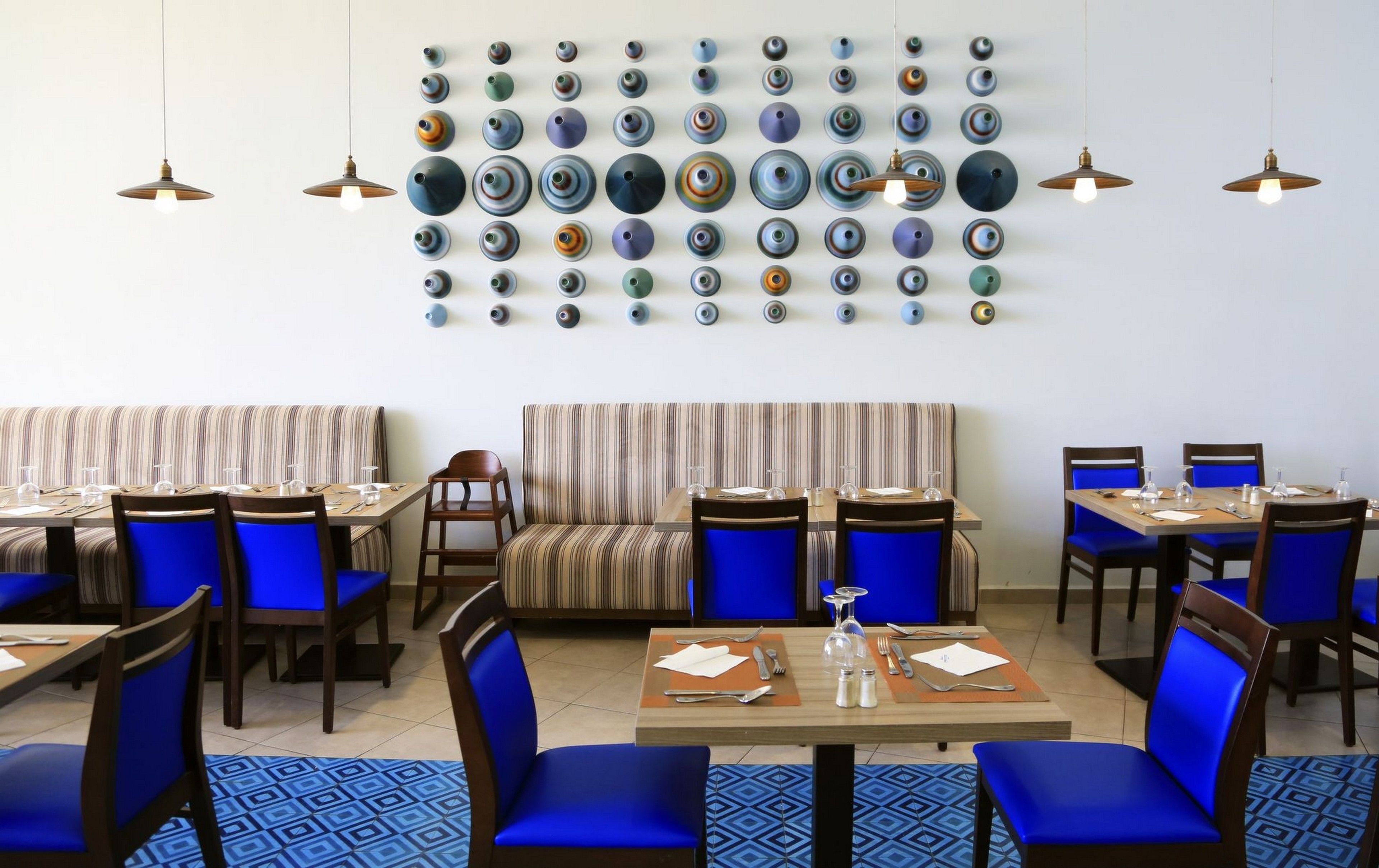 Un restaurant avec des banquettes et des tables à rayures, des chaises bleues et un mur décoré d’assiettes.