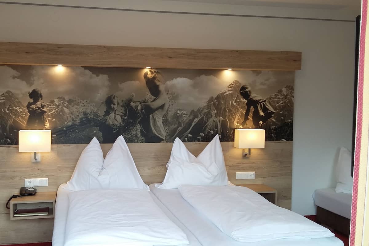 Habitación de hotel con una cama grande, dos lámparas de noche, una mesita de noche y un mural en la pared con un paisaje montañoso.