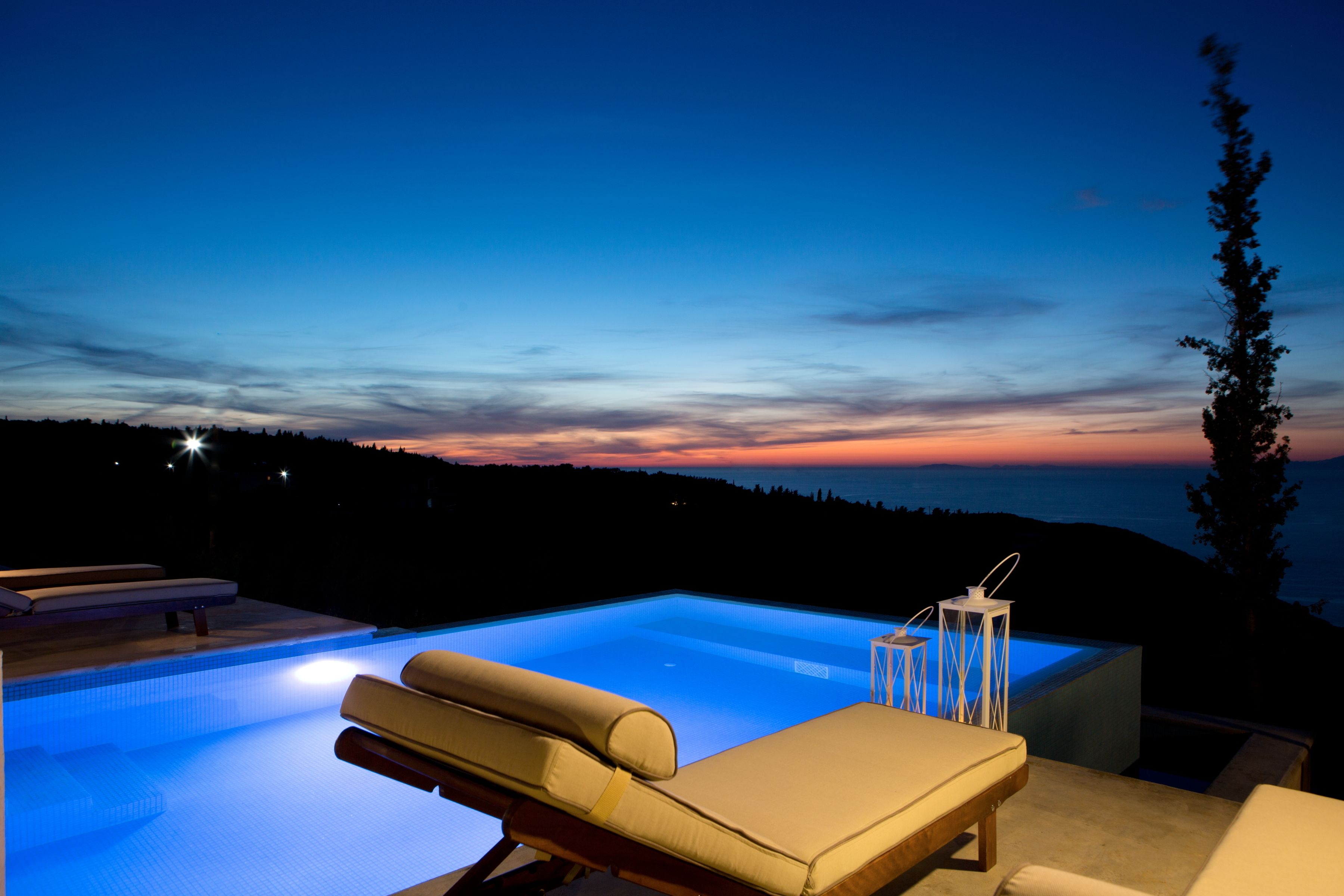 A swimming pool with lounge chairs and a view of the sunset over a forested landscape.