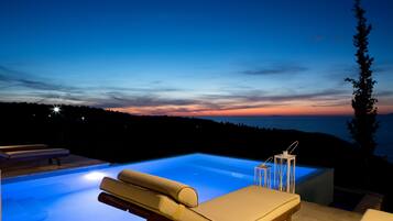 A swimming pool with lounge chairs and a view of the sunset over a forested landscape.