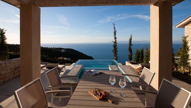A wooden deck with a pool, lounge chairs, and a table with wine glasses and fruit.