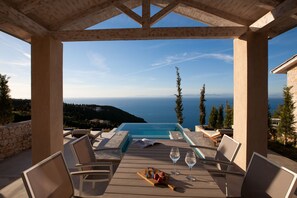 A wooden deck with a pool, lounge chairs, and a table with wine glasses and fruit. - Ionian Horizon Villas (Lefkada)