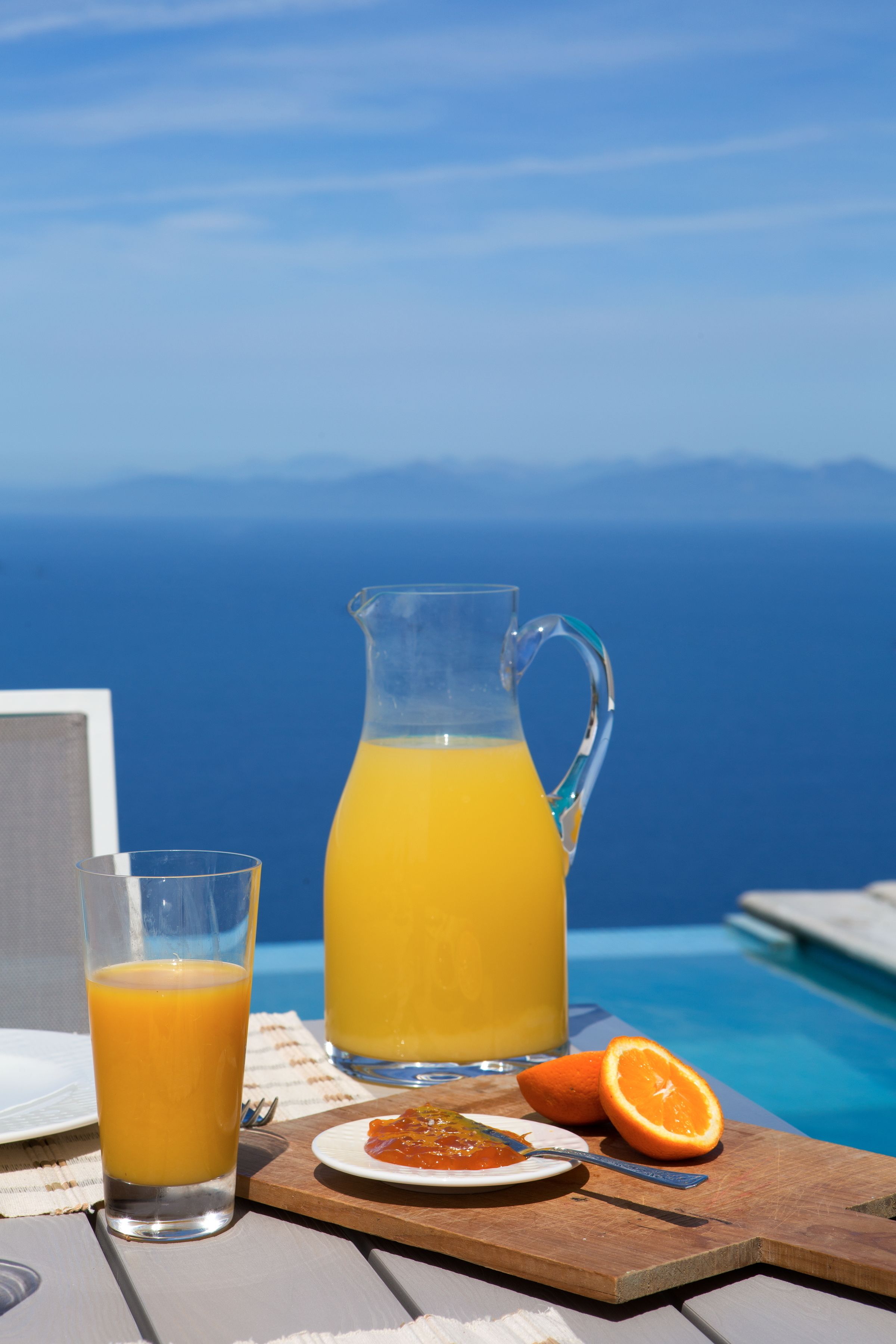 A table with a glass of orange juice, a pitcher of orange juice, a plate with a slice of orange, and a small dish of jam, set against a poolside view.