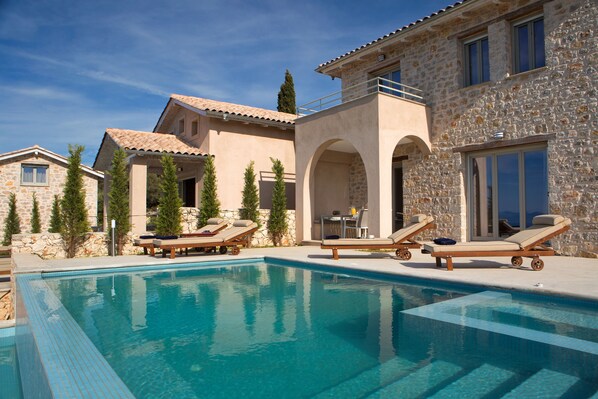 A pool area with lounge chairs and a stone building with arched windows. - Ionian Horizon Villas (Lefkada)
