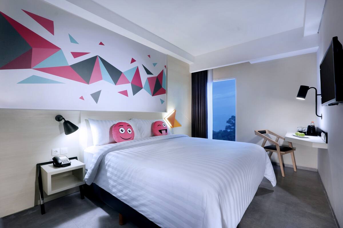 Ein modernes Hotelzimmer mit einem großen Bett, einem Schreibtisch mit Stuhl, einem Flachbildfernseher und einer Wand mit geometrischen Formen.