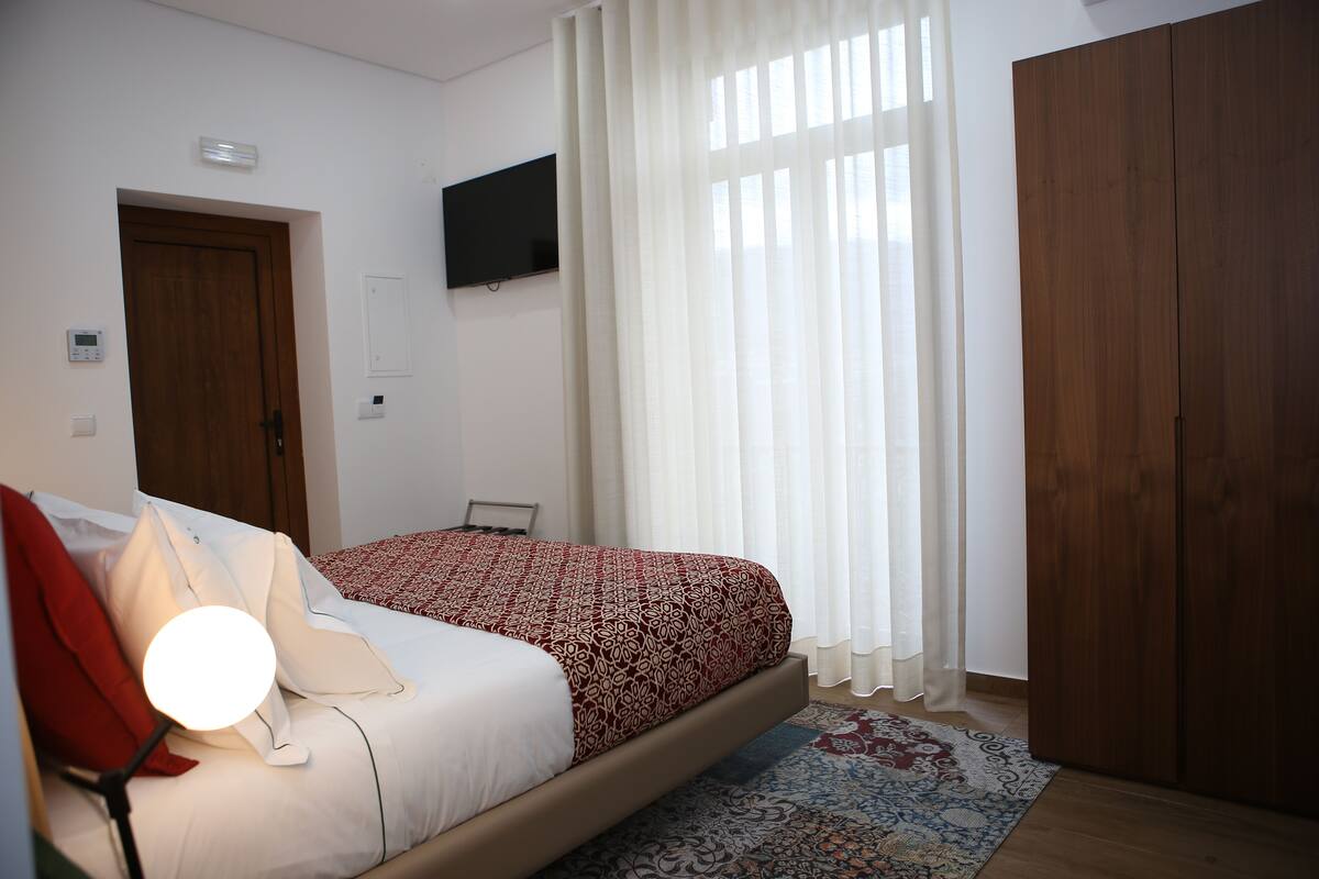 Ein Schlafzimmer mit einem Bett, einem an der Wand montierten Fernseher, einem Holzschrank und einem Fenster mit Vorhängen.