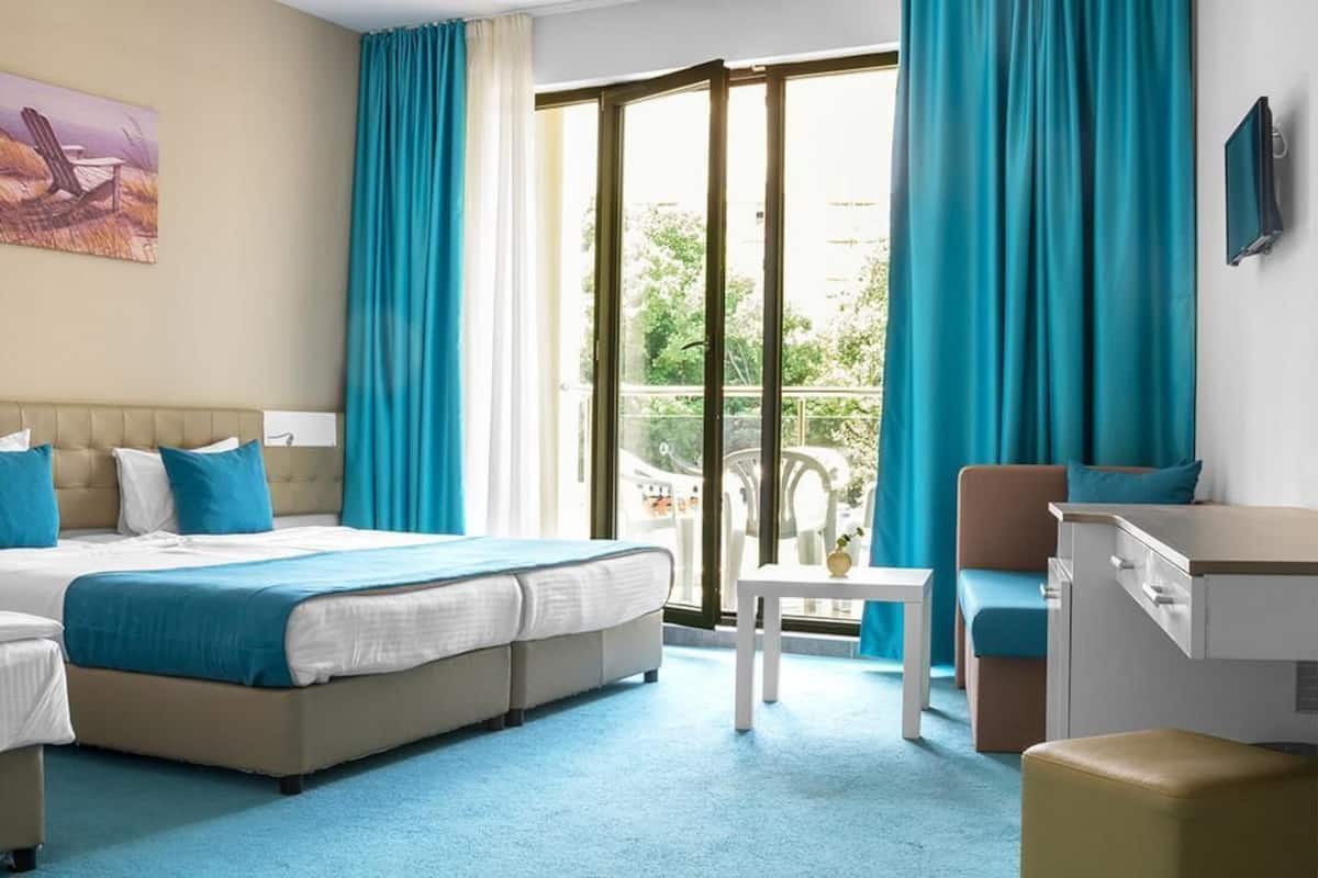 A hotel room with a large bed, a desk, a chair, and a balcony with curtains.
